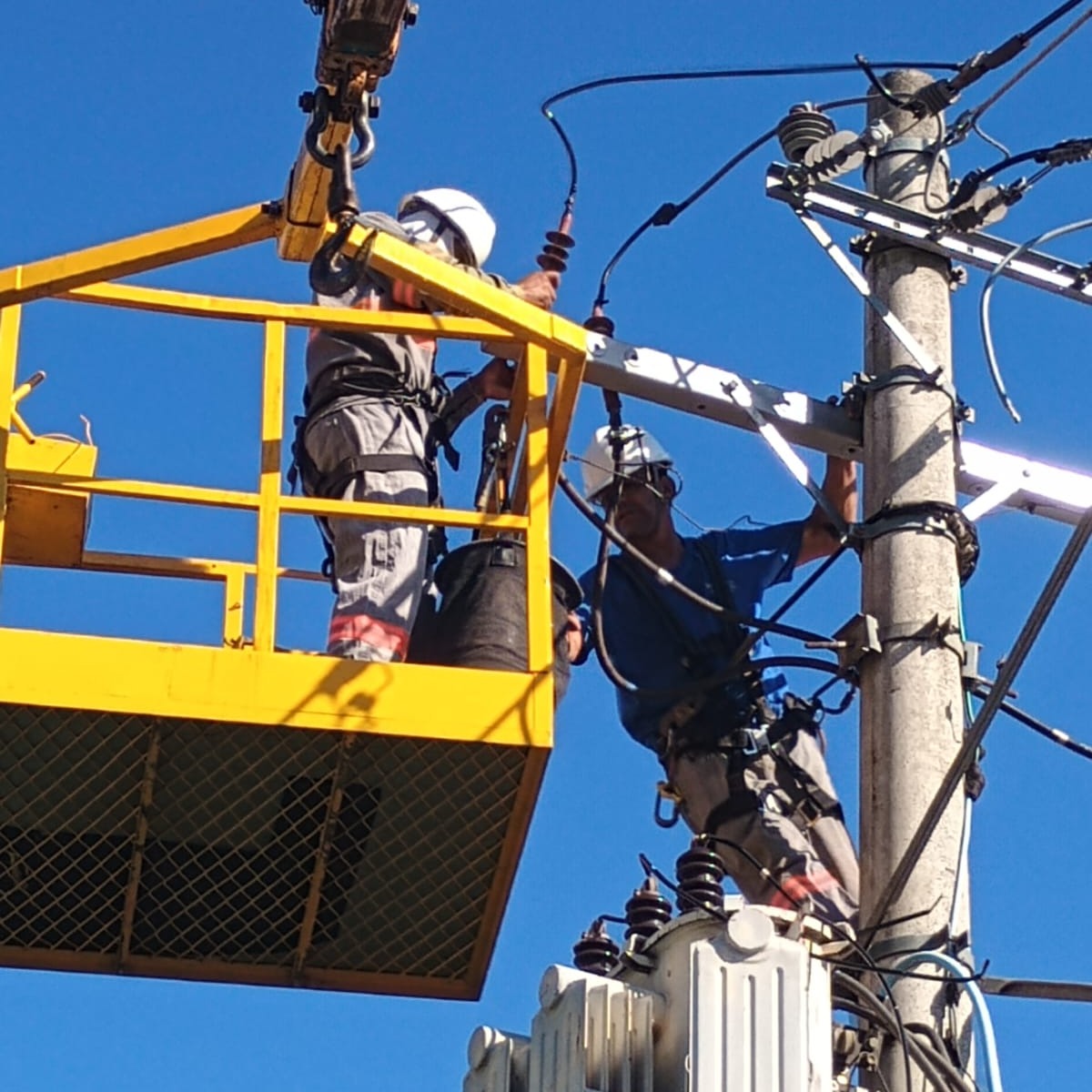 Dois técnicos com capacete e cinto de segurança trabalhando no topo de um poste, um na cesta do munck e outro escalado na estrutura